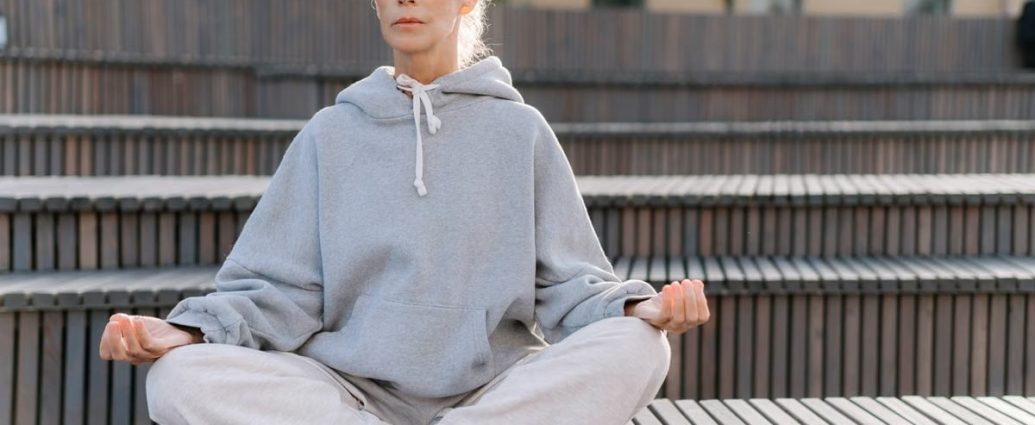 Elderly woman in gray hoodie meditating outdoors, promoting fitness and relaxation.