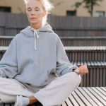 Elderly woman in gray hoodie meditating outdoors, promoting fitness and relaxation.