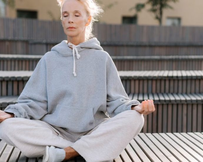 Elderly woman in gray hoodie meditating outdoors, promoting fitness and relaxation.