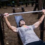 Man weightlifting outdoors, using a bench press setup amid trees for strength training.