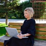 Woman sitting on a bench with a laptop, enjoying a sunny day in a green park setting.